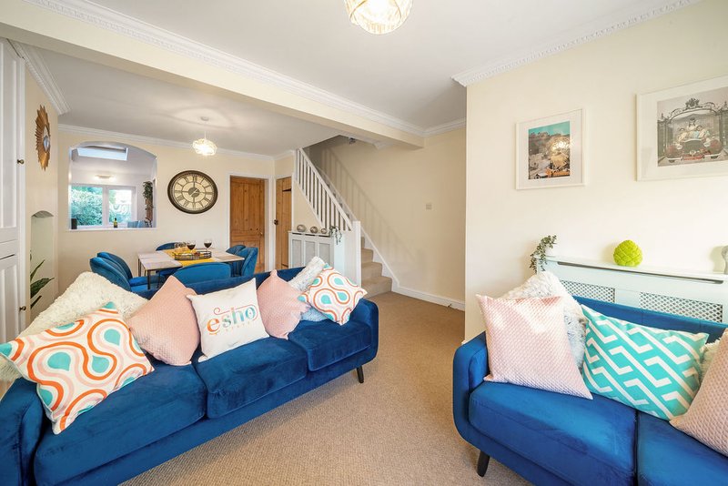 Open-plan living area with two blue sofas, colorful cushions, stairs, and wall art