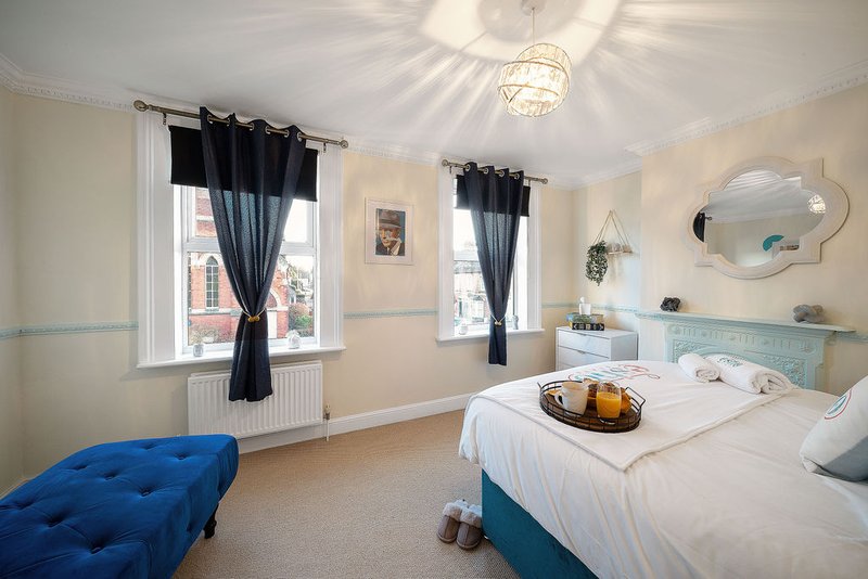 Bright bedroom with double bed, blue ottoman, dual windows with black curtains and decorative mirror