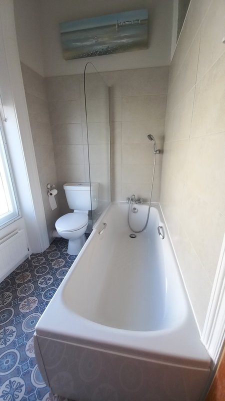 Bathroom with white bathtub, toilet, and window with coastal artwork above