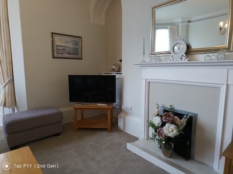 Living room with fireplace, TV on wooden stand, grey upholstered ottoman, and mirror above mantelpiece