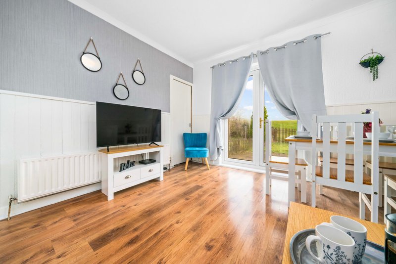 Bright open-plan living room with wooden flooring, TV unit, blue armchair, cot, and glass doors overlooking garden