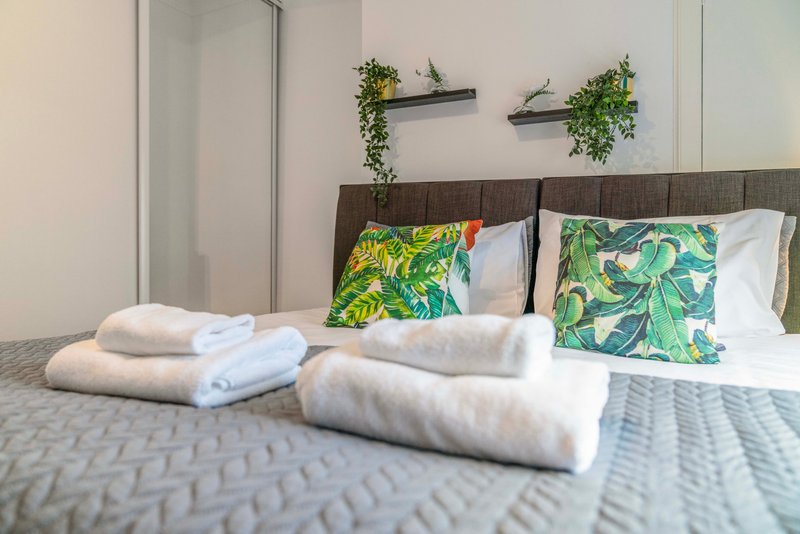 Bedroom with brown headboard, white rolled towels, tropical patterned cushions, and potted plants on shelves