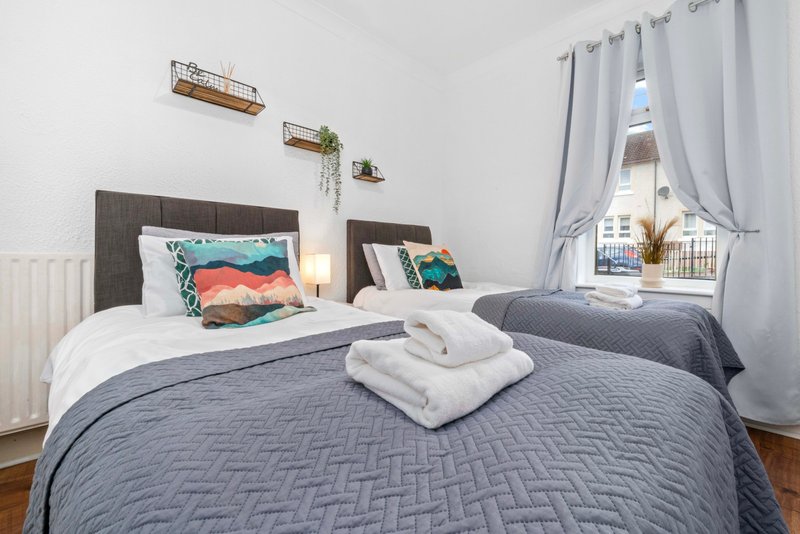 Bright bedroom with two beds, gray quilts, white pillows, dark headboards, and window with curtains