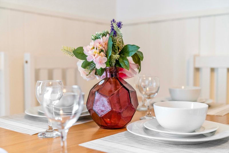 Dining table set with white dinnerware, glassware, and red vase with pink and blue flowers