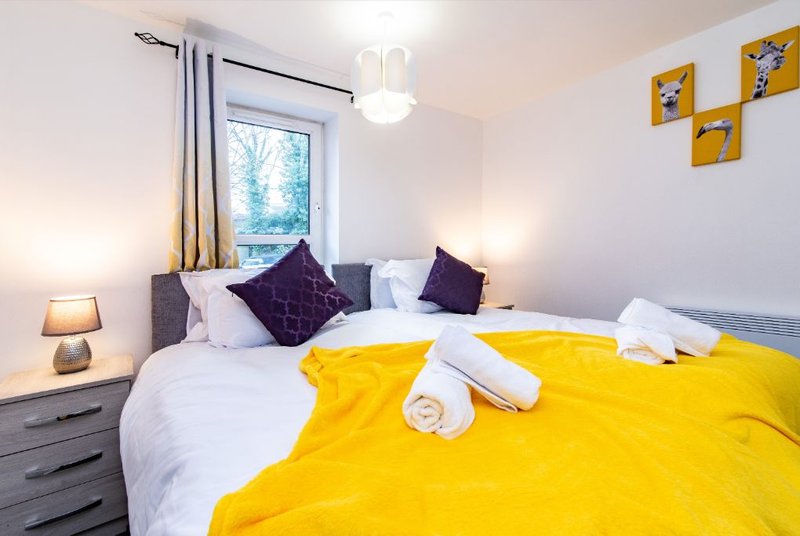Bright bedroom with yellow bedding, purple cushions, white pillows, bedside lamp and window view