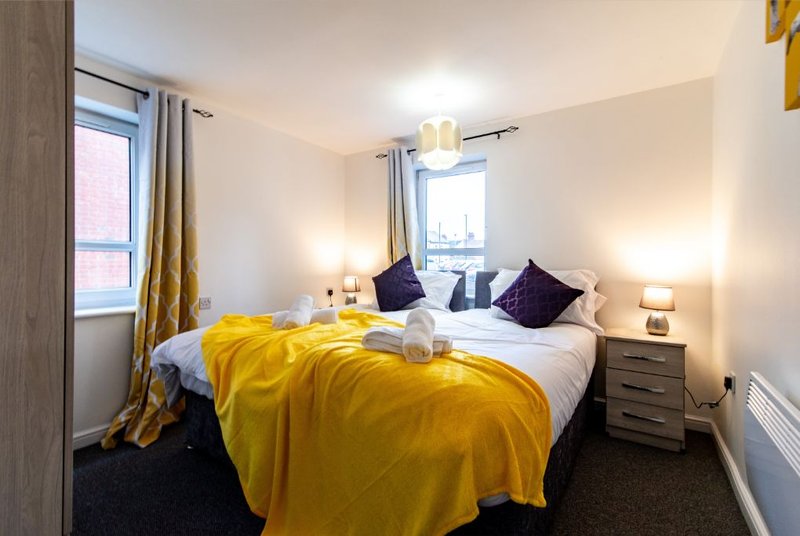 Bright bedroom with double bed, yellow throw, purple cushions, bedside lamps and windows