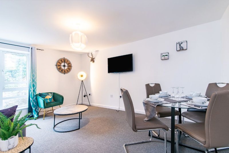 Open-plan living and dining area with turquoise armchair, wooden coffee table, wall-mounted TV, and dining table with chairs