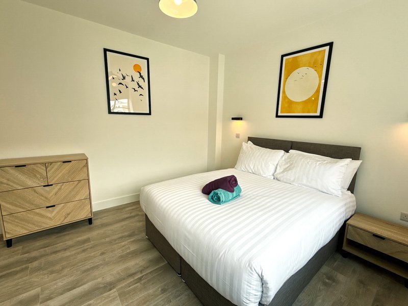 Modern bedroom with queen bed, white bedding, wooden dresser, framed wall art and wooden flooring