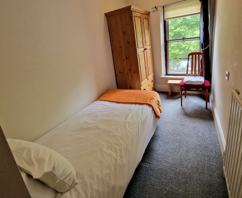 Narrow bedroom with single bed, wooden wardrobe, chair by window with garden view
