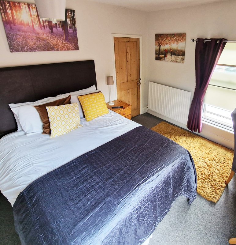 Bright bedroom with double bed, black headboard, patterned cushions, wooden door and purple curtains
