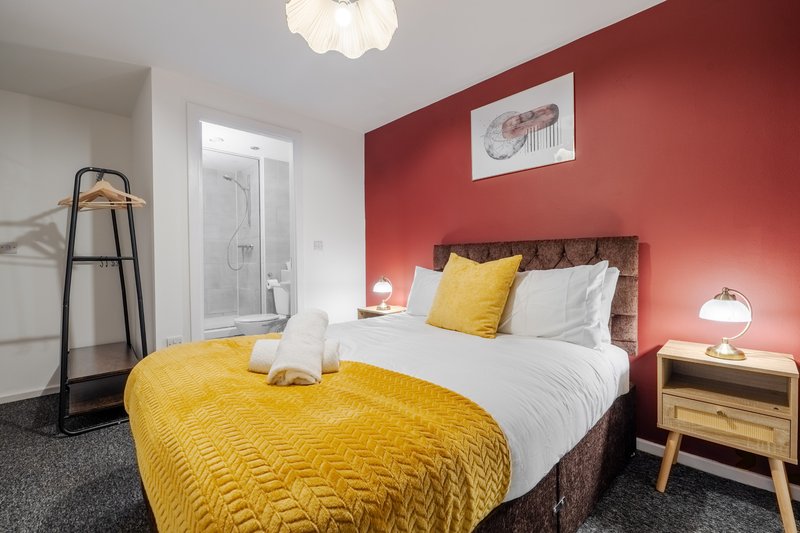 Modern bedroom with double bed, yellow bedding, red accent wall, wooden nightstand and ensuite bathroom