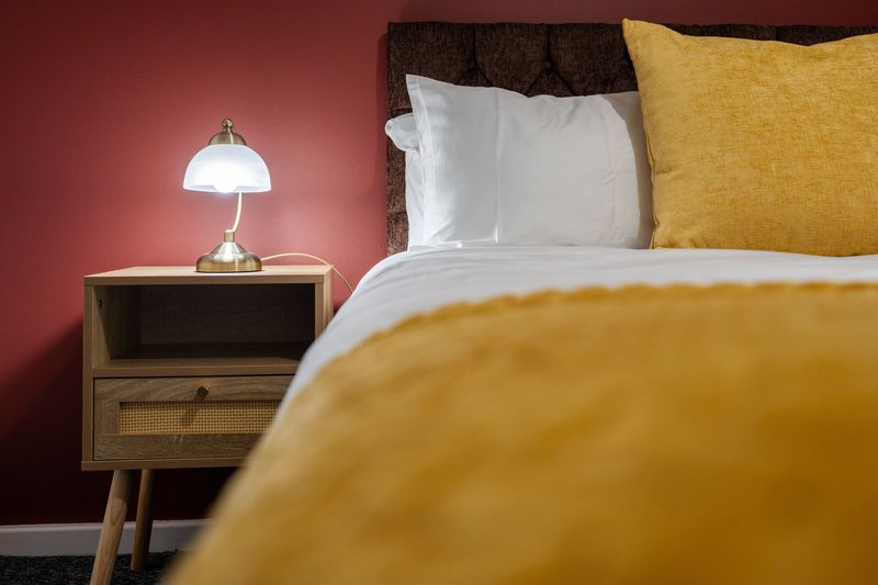 Modern bedroom with mustard yellow bedding, white pillows, wooden nightstand and table lamp against pink wall