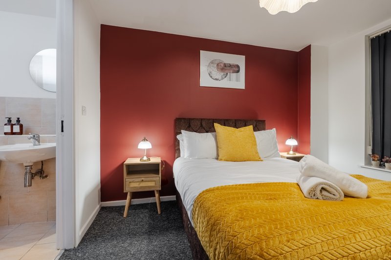 Modern bedroom with double bed, yellow throw, red accent wall and ensuite bathroom visible
