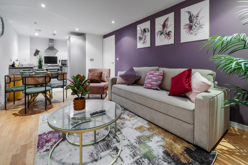 Open-plan living room with purple accent wall, grey sofa, round glass table, and kitchen area