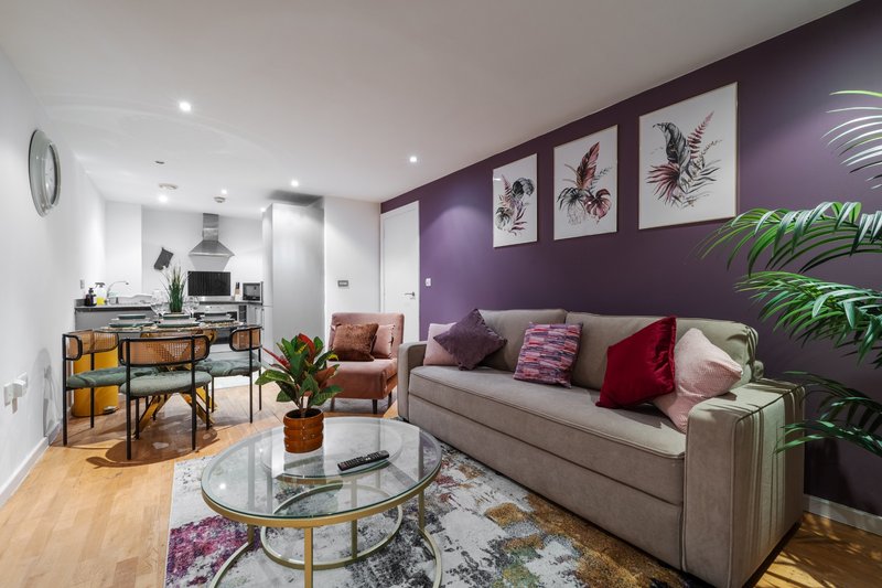 Open-plan living room with purple accent wall, grey sofa, round glass table, kitchen area, and potted plants