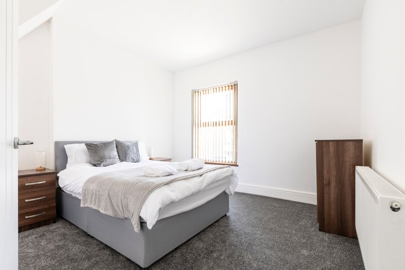 Bright bedroom with grey bed, wooden nightstand, vertical blinds and grey carpet flooring
