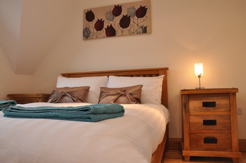 Bedroom with wooden bed frame, white bedding, teal throw blanket, and wooden nightstand with lamp