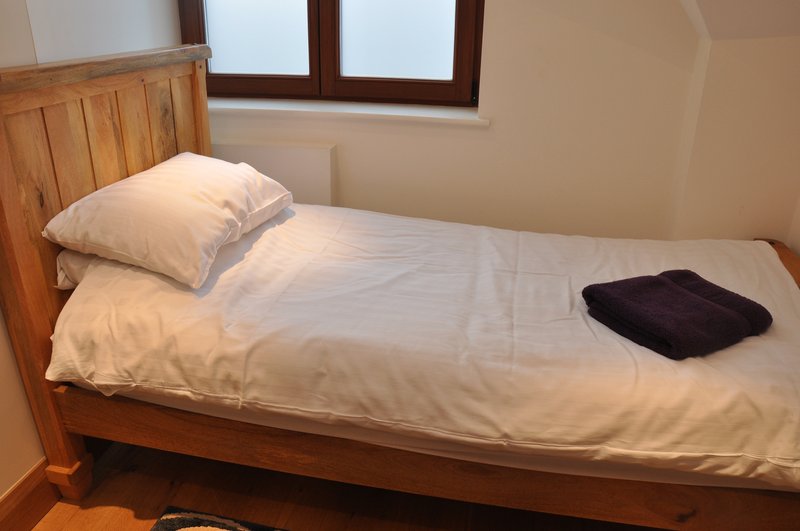 Single wooden bed with white bedding, cream pillow, and black garment on mattress
