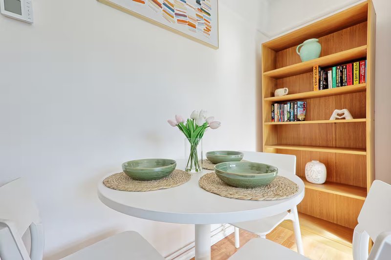 Bright dining area with round white table, green bowls, flowers and wooden bookshelf
