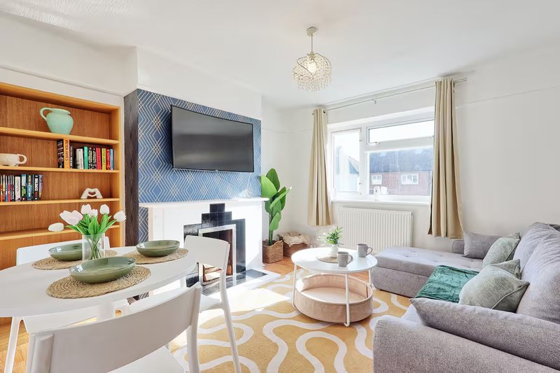 Bright modern living room with grey sofa, wall-mounted TV, wooden bookshelf, dining table and large windows