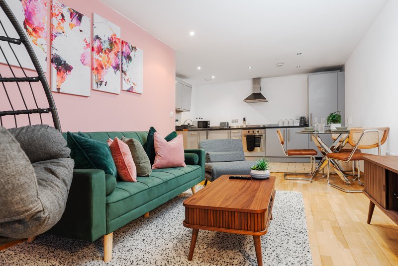 Open-plan living space with green sofa, wooden coffee table, pink accent wall, and kitchen area