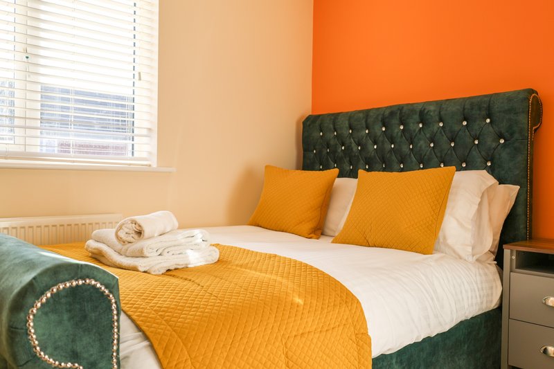 Bedroom with double bed, green headboard, orange and yellow bedding, rolled towels