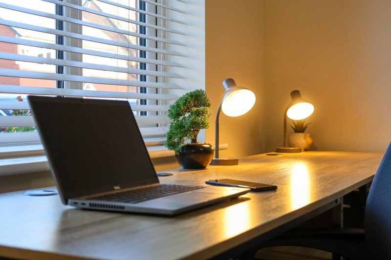 Modern desk workspace with laptop, desk lamps, plant, and city view through blinds