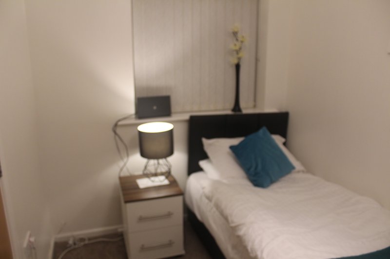 Small bedroom with single bed, white bedding, blue cushion, wooden nightstand and lamp