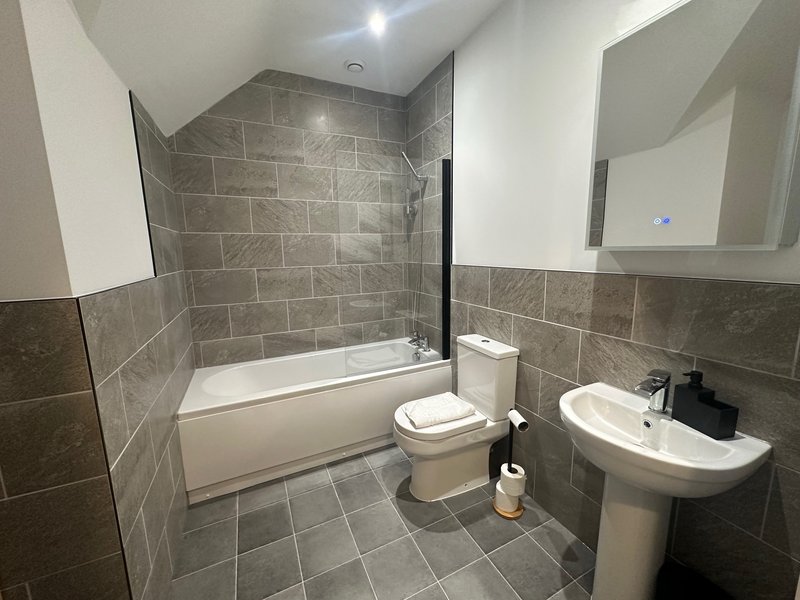 Modern bathroom with grey tile walls, bathtub, toilet, pedestal sink and mirror