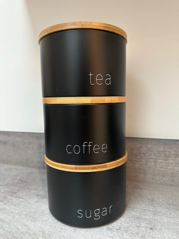 Black cylindrical kitchen storage container with wooden lid and brass bands, labeled tea, coffee, and sugar