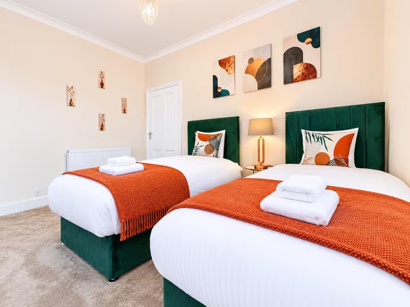 Modern twin bedroom with green upholstered headboards, orange bedding, wall art, and ceiling pendant light