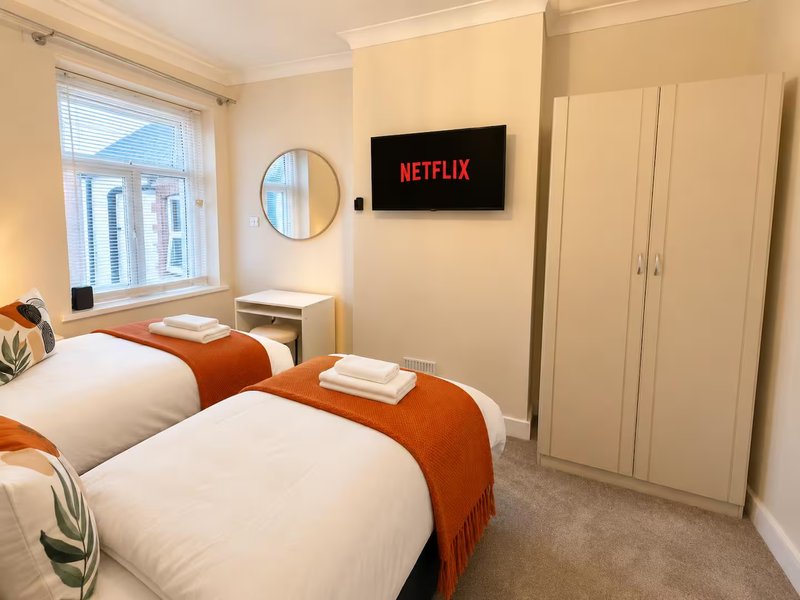 Twin bedroom with two single beds, orange throws, wall-mounted TV, wardrobe and window with blinds
