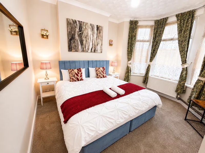 Bright bedroom with blue upholstered bed, white bedding, red runner, and olive curtains