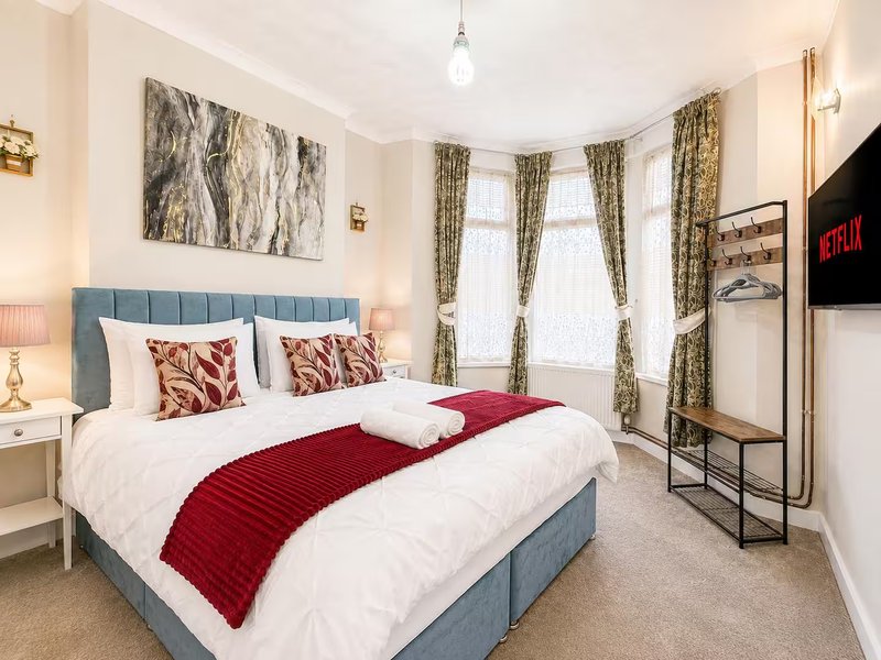 Bright bedroom with blue upholstered bed, red bedding, bay windows with patterned curtains, and wall-mounted TV