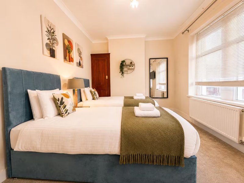 Bright bedroom with double bed, blue headboard, olive throw, framed wall art and window