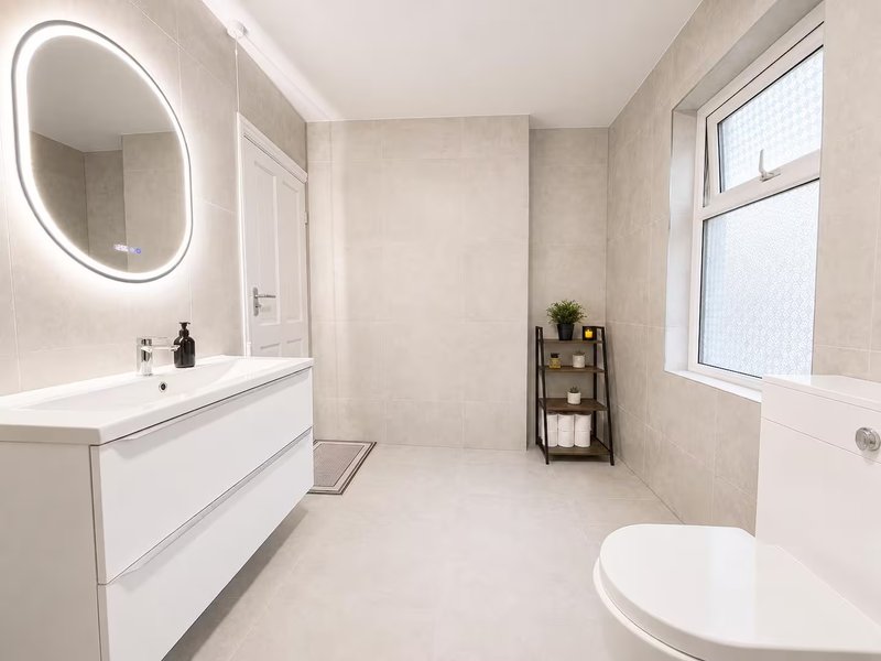 Modern bathroom with illuminated round mirror, white vanity, toilet, and storage shelving