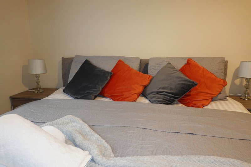Bedroom with grey upholstered bed, orange and black cushions, white bedding and table lamps