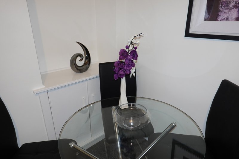Round glass dining table with purple flowers in vase, metal sculpture and framed art on wall