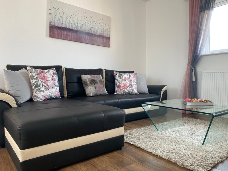 Modern living room with black and cream L-shaped sofa, decorative floral cushions, glass coffee table, and window with curtains.