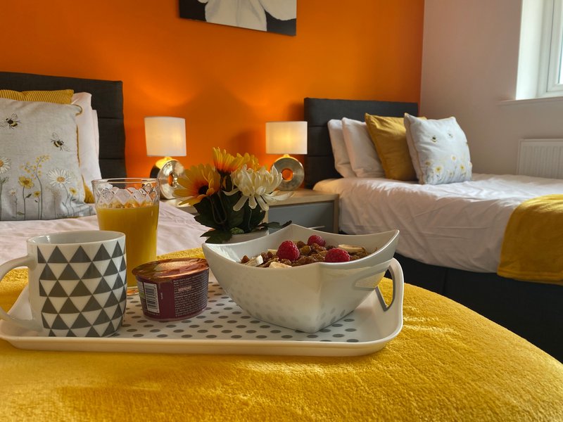 Bright bedroom with two beds, orange walls, yellow bedding, and decorative table with flowers and mugs
