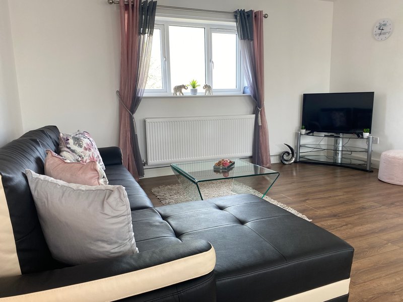 Living room with black leather sofa, cushions, glass coffee table, TV, and window with curtains