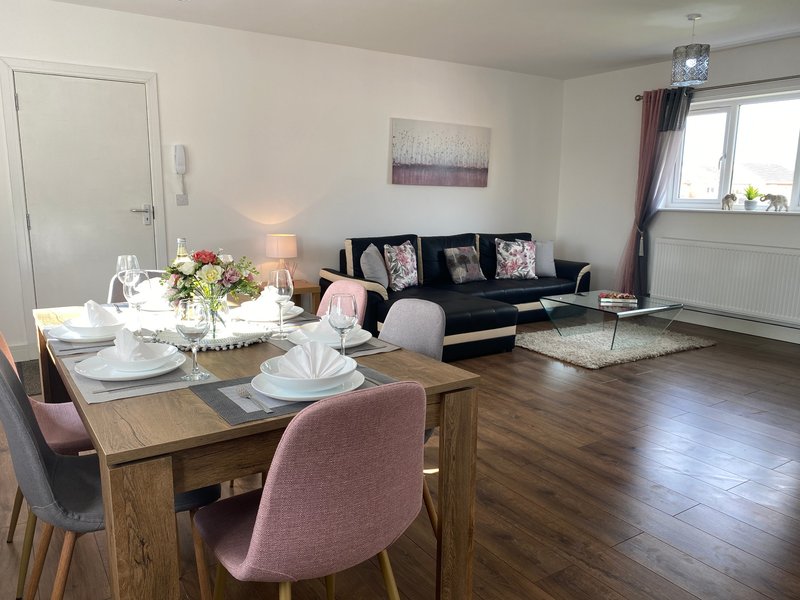 Open-plan living and dining area with wooden table, pink chairs, black sofa, and window with curtains