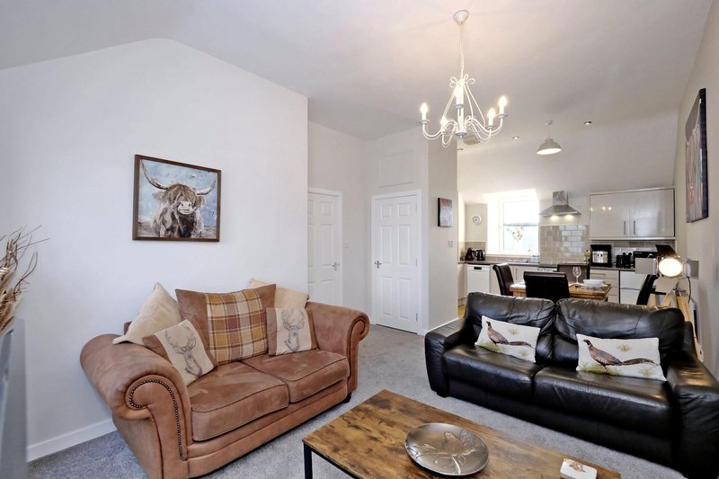 Open-plan living room with brown leather sofa, black sofa, chandelier, and kitchen visible in background