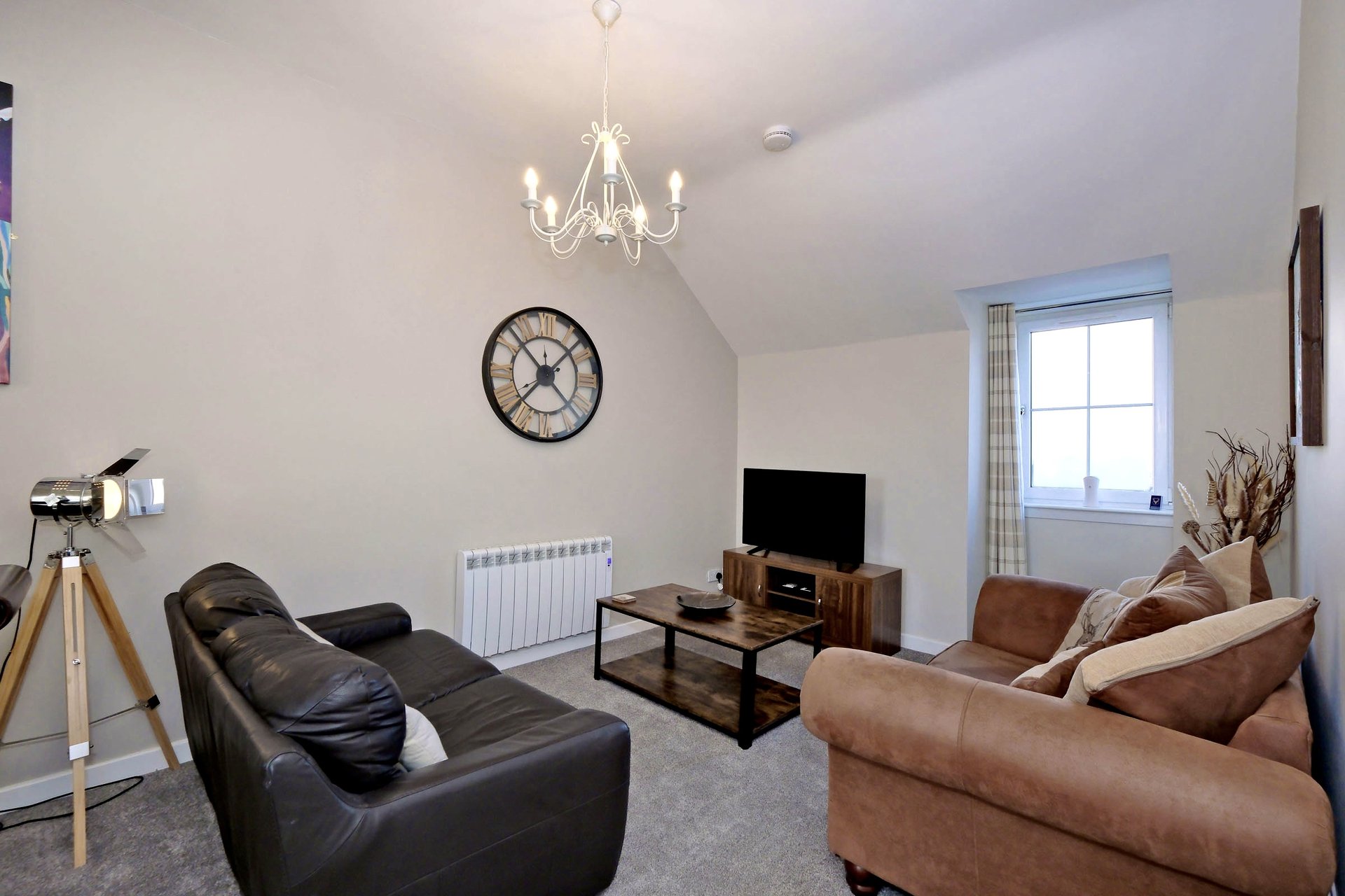 Bright living room with black leather armchair, tan sofa, TV on wooden stand, chandelier and wall clock