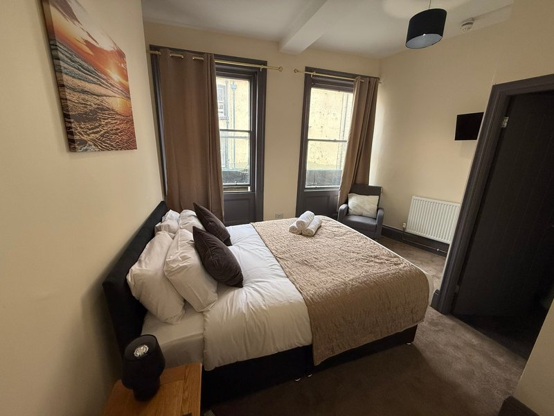 Bedroom with double bed, beige bedding, dark headboard, two windows with curtains, and cream walls