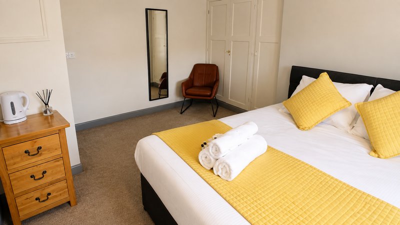 Bedroom with double bed, yellow bedding and pillows, wooden nightstand, mirror and armchair
