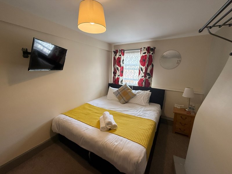 Bedroom with double bed, yellow throw, wall-mounted TV, window with red curtains