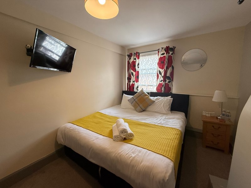 Bedroom with double bed, yellow throw, wall-mounted TV, window with red curtains