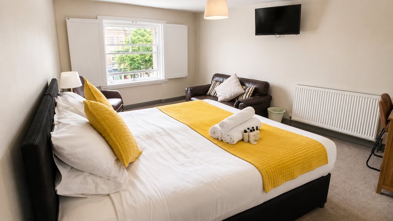 Spacious bedroom with double bed, yellow throw cushions, TV wall mount, seating area and window.