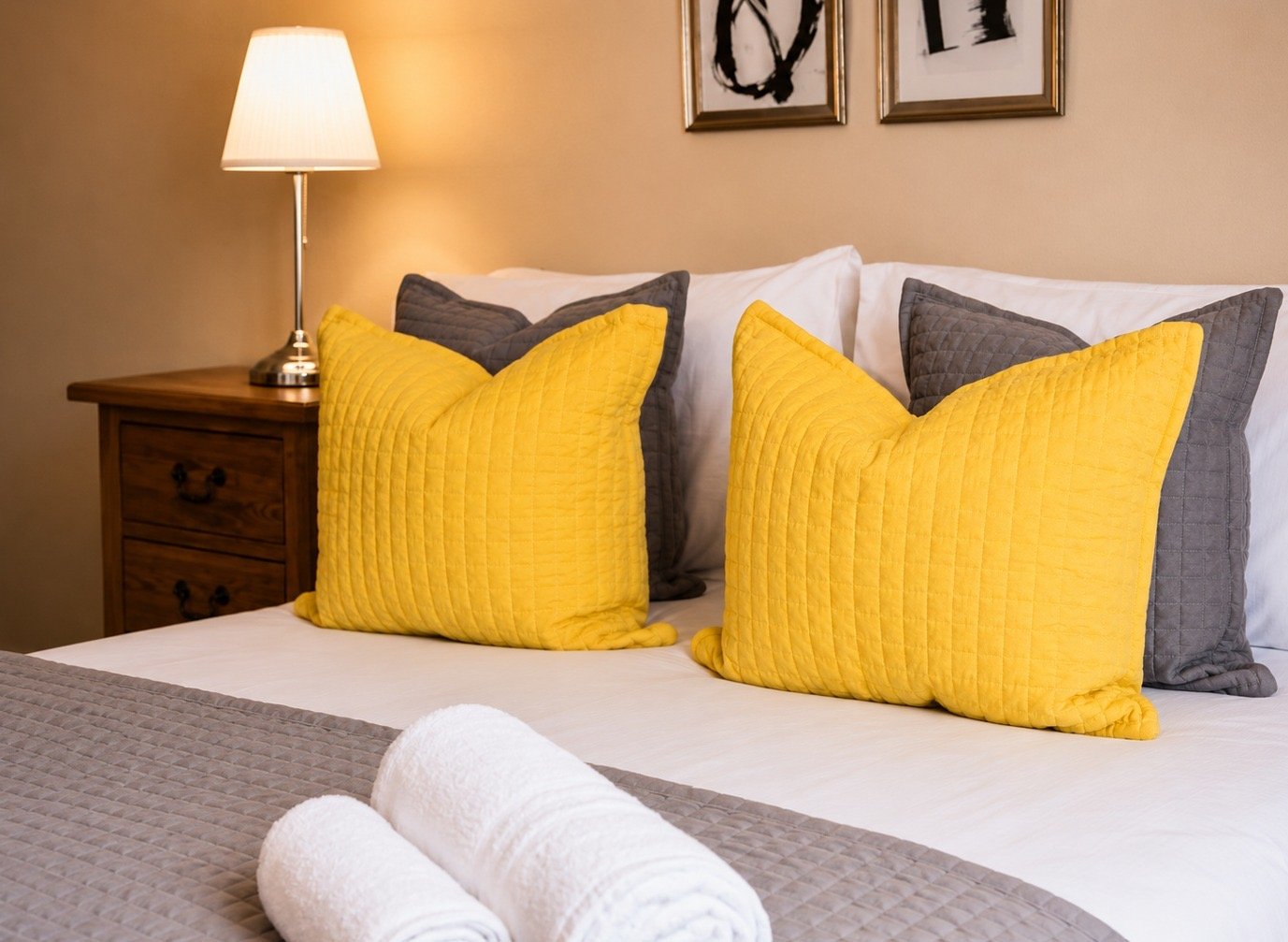 Bedroom with bed, yellow and grey cushions, wooden nightstand and lamp